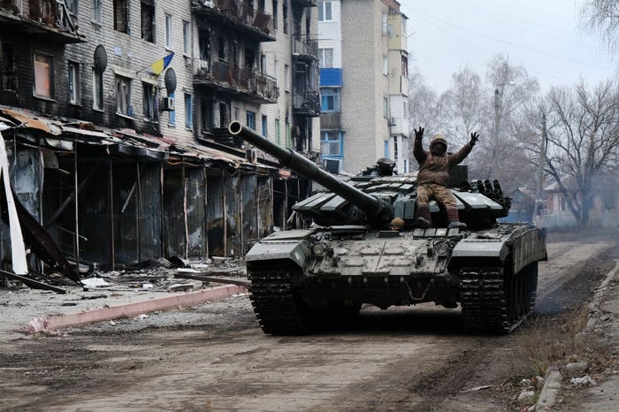 A person on a military tank makes peace signs as it drives down a dirt road past heavily damaged buildings with a Ukrainian flag visible.