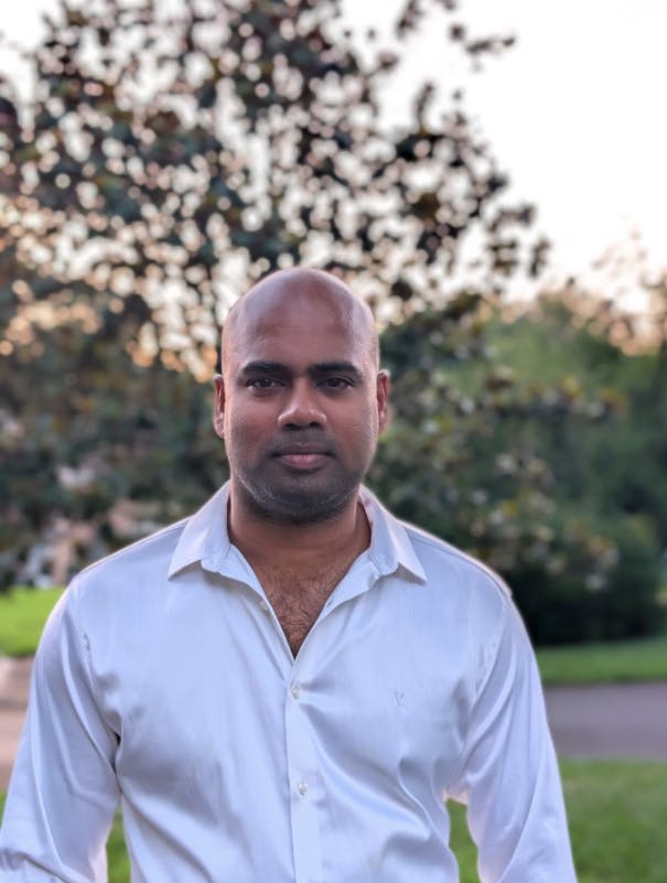 "Man with a bald head and light stubble wearing a white collared shirt, standing outdoors with blurred trees in the background."