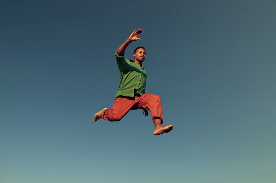 Man in a green shirt and orange pants leaping in the air against a clear blue sky.