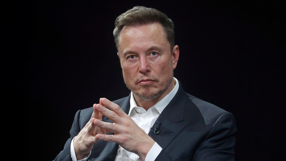 "Elon Musk in a dark suit and white shirt, with his hands clasped, looking forward against a dark background."