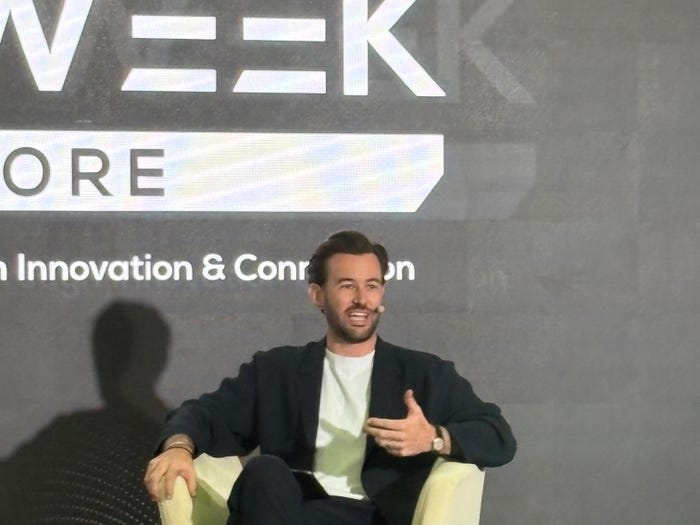 Man speaking and gesturing from an armchair, with event branding 'WEEK ORE' and 'Innovation & Con' on the dark background.