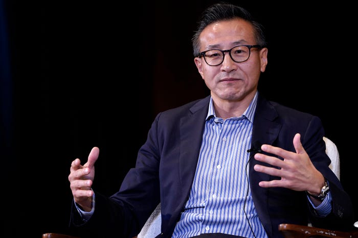 Man in glasses, striped shirt, and dark blazer gesturing with open hands while speaking.