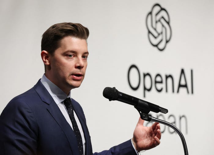 Man in a suit and tie speaking into a microphone, with the OpenAI logo and text clearly visible in the background.