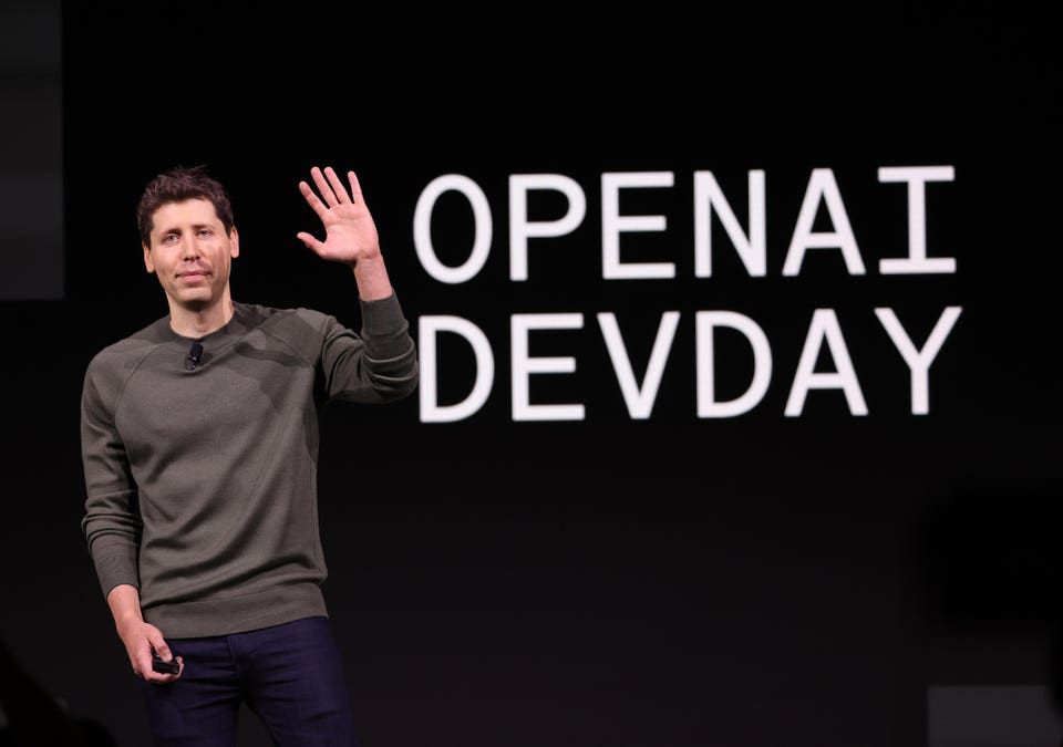 Sam Altman on stage at OpenAI DevDay, raising his hand, with "OPENAI DEV DAY" displayed behind him.