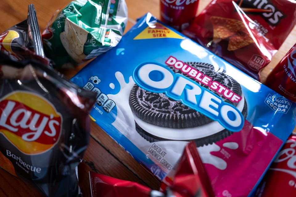 Double Stuf Oreo cookies bag, with bags of Lay's Barbecue chips, Doritos, Tostitos, and Coca-Cola cans in the background.