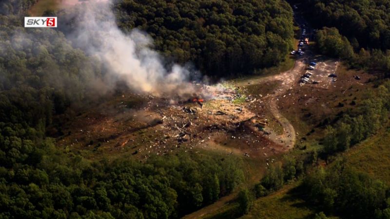 Aerial view of a plane crash site with widespread debris, smoke, and flames in a clearing, surrounded by forest. Vehicles are visible on a dirt path and in a parking area.