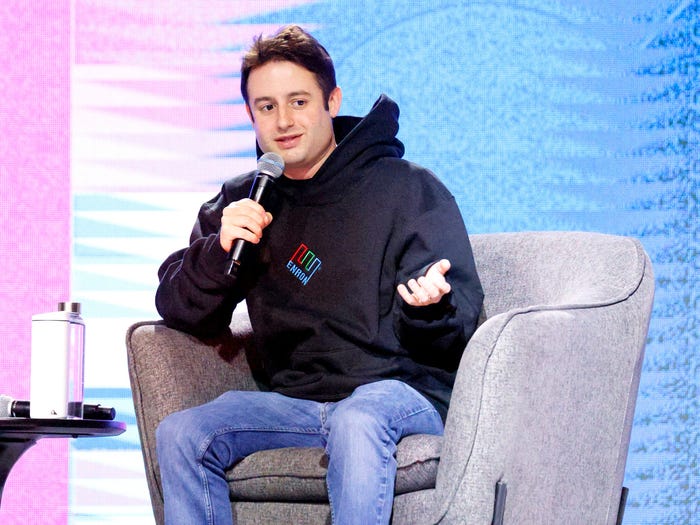 Man in a black "ENRON" hoodie and jeans speaking into a microphone while sitting in a grey armchair.