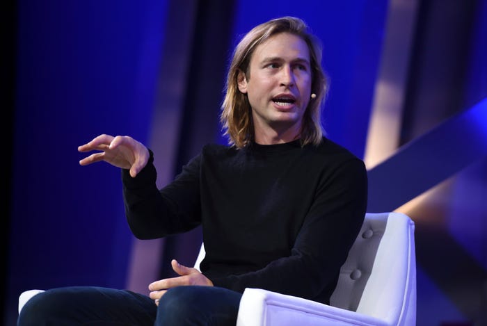 Blonde-haired man in a black shirt speaking and gesturing, seated in front of a blue background.