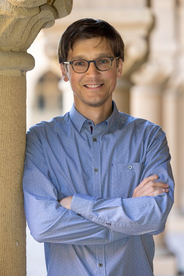Smiling man in glasses and a blue patterned shirt, with arms crossed, leaning against a stone column.