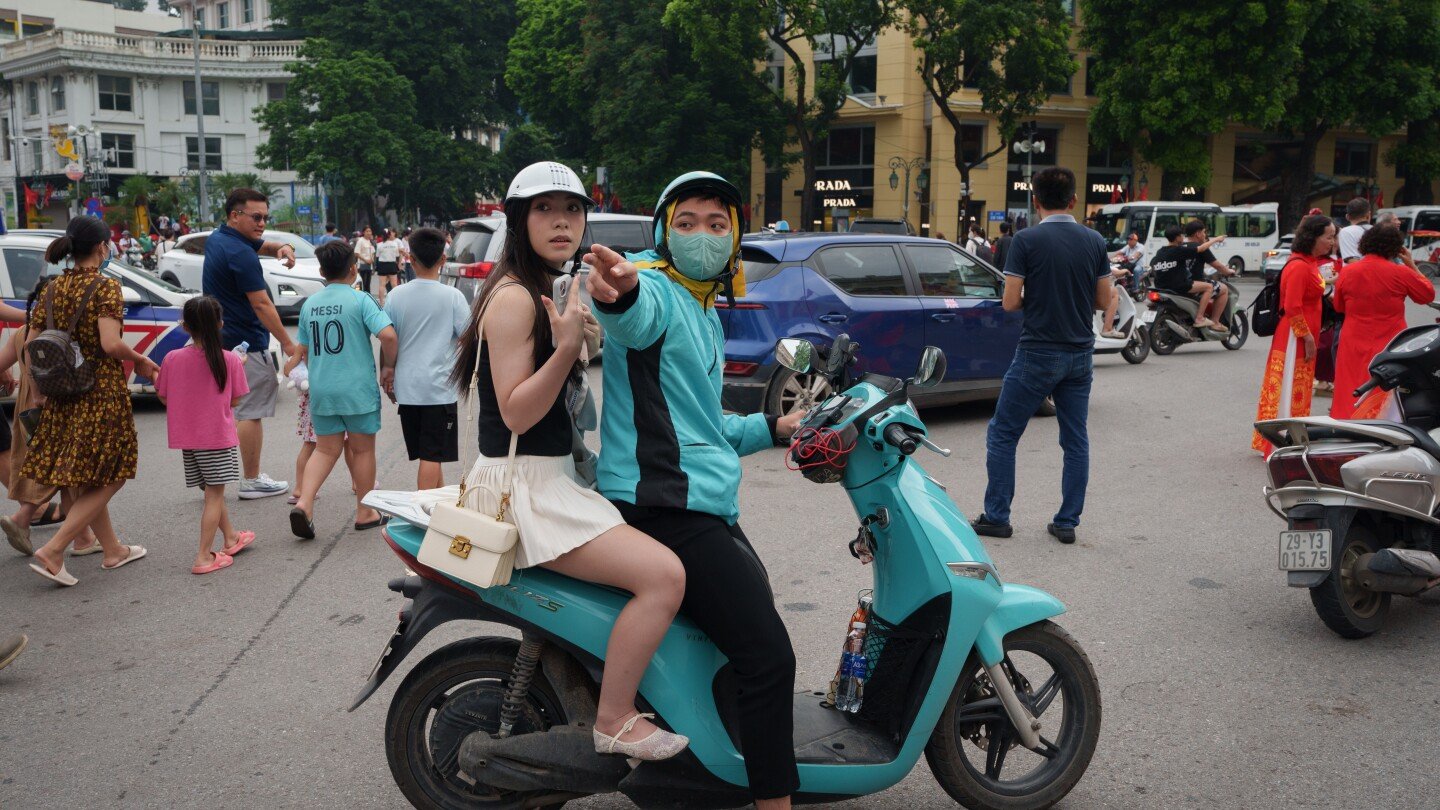 A man and woman on a teal scooter on a busy city street. The masked man, in a blue jacket, points forward while the woman, in a white helmet and skirt, looks wide-eyed to the side.