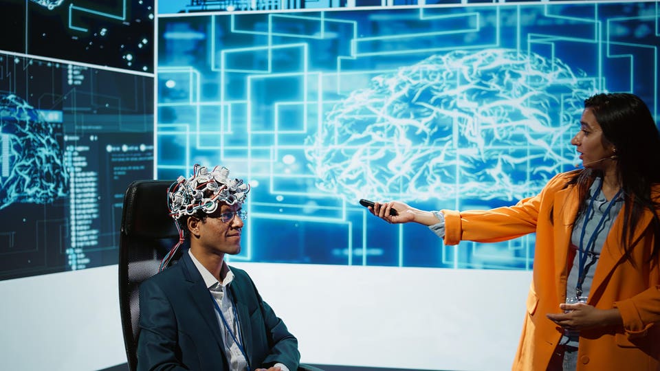 Man wearing an EEG cap and glasses sits facing a woman holding a remote control, with large screens displaying a glowing brain diagram and data in the background.