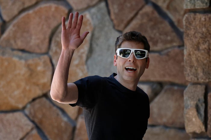 Man in white sunglasses and a black t-shirt waving with a wide smile against a stone wall background.