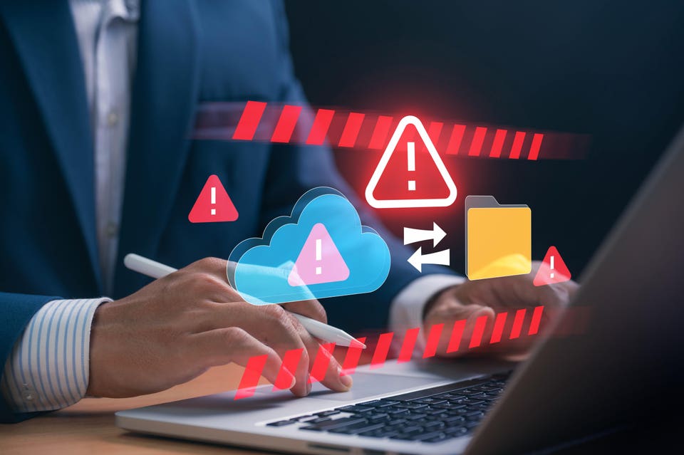 Hands using a laptop with holographic red warning signs, a cloud storage icon showing an alert, and a file folder, depicting a data security breach or system failure.