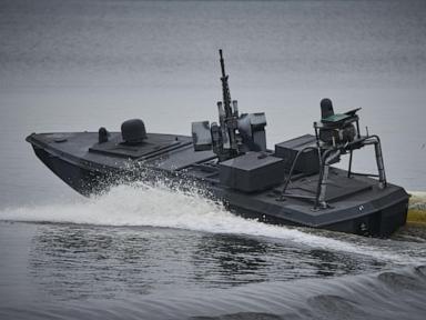 Dark military patrol boat with mounted machine guns moving across water, creating a wake.