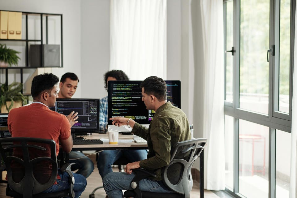 Four men collaborating on computer code in an office, with two men pointing at screens showing lines of code.