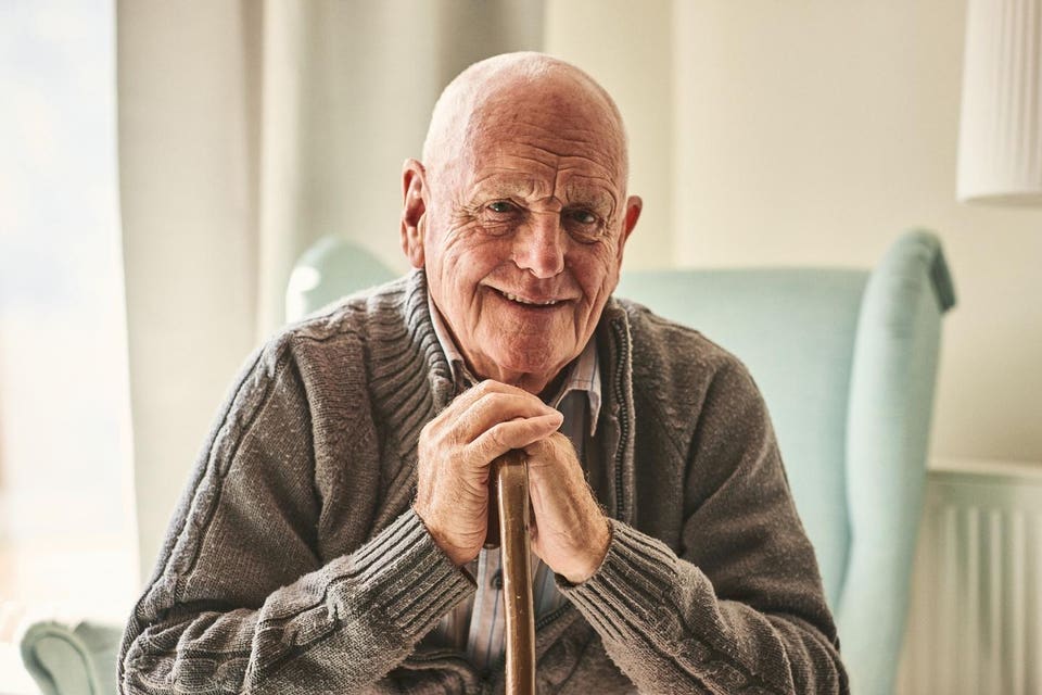 Smiling elderly man in a grey cardigan, sitting in an armchair with hands clasped over a walking stick.