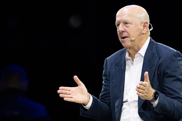 Bald man in a blue suit and white shirt, wearing a headset microphone, speaking and gesturing with hands.
