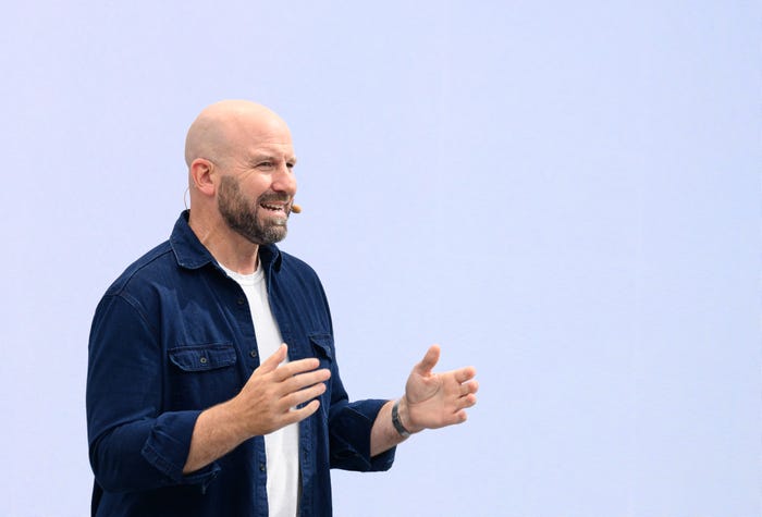 "A bald, bearded man in a blue shirt with a microphone, gesturing as he speaks."