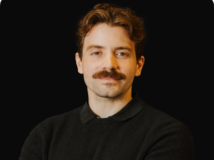 "Man with brown hair and a prominent mustache, wearing a black collared sweater, looking directly at the viewer against a black background."