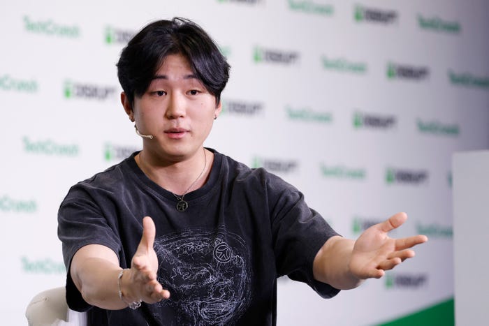 East Asian man speaking and gesturing with open hands, wearing a dark t-shirt and an ear-worn microphone, against a background with green logos.