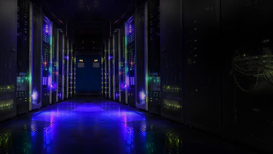 Long aisle in a data center with server racks glowing with blue, green, and purple lights, reflecting on the floor.