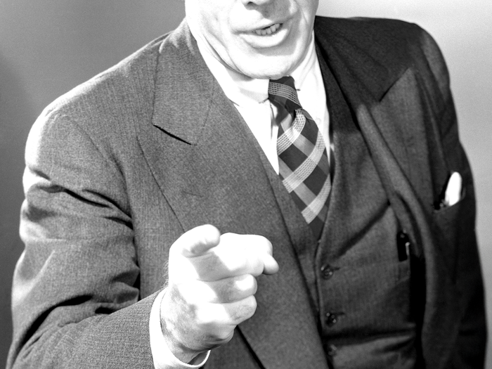 Man in a suit, tie, and waistcoat pointing directly at the viewer with an open mouth.