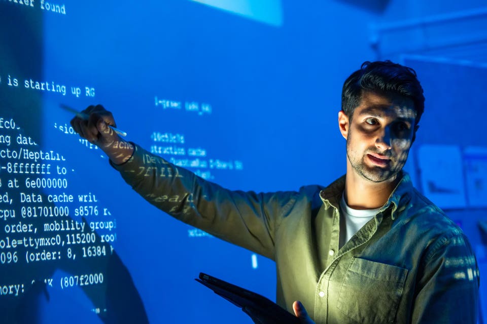 A man points a pen at a large blue screen displaying white technical information. The screen's light and projected text illuminate his face and arm as he holds a tablet.